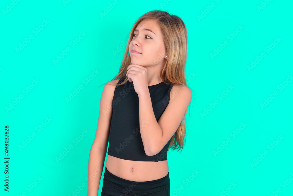 Thoughtful blonde little kid girl wearing black sport clothes over green background holds chin and looks away pensively makes up great plan Thoughtful blonde little kid girl wearing black sport clothes over green background holds chin and looks away pensively makes up great plan