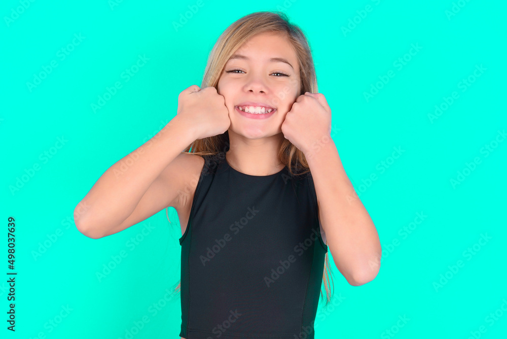 Fototapeta premium Happy blonde little kid girl wearing black sport clothes over green background keeps fists on cheeks smiles broadly and has positive expression being in good mood
