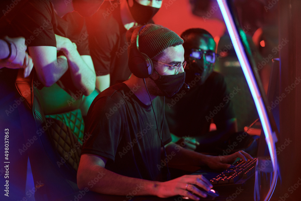 Concentrated young Arabian gamer in hat and cloth mask playing video game while his team members watching him