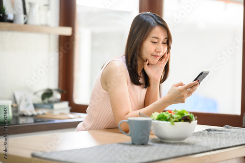 Happy smiling Asian woman using smartphone mobile at home in kitchen with salad bowl on the table