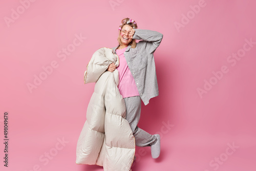 Energetic cheerful woman wears slumber suit and slippers makes hair curly applies rollers holds soft blanket isolated over pink background going to have healthy sleep. People and rest concept