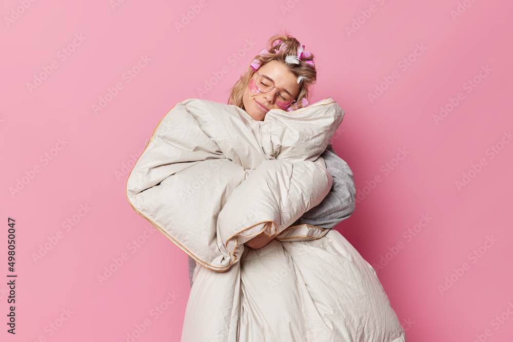Pleased young European woman sees sweet dreams tilts head holds blanket prepares for party makes hairstyle applies hair rollers and beauty patches isolated over pink bakground. Time to have rest