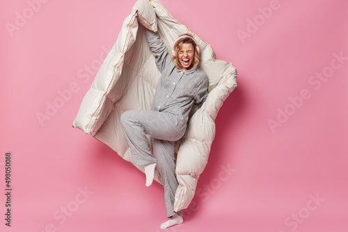 Joyful energetic young woman laughs out gladfully listens music via headphones enjoys favorite playlist dressed in nightwear poses with blanket awakes in good mood dances against pink background.