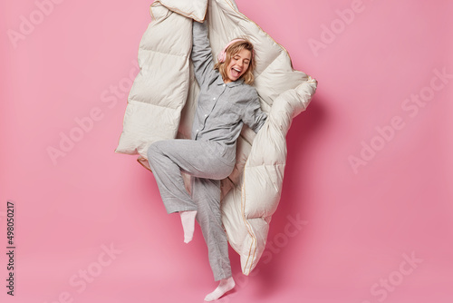 Glad happy woman dances and has fun listens music via wireless headphones holds blanket foolishes around feels energetic after healthy sleep isolated over pink background. Wakeing up concept