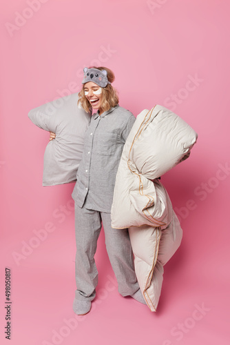 Positive young European woman dressed in grey nightwear holds pillow and blanket laughs joyfully applies beauty patches under eyes goes to bed isolated over pink background. Full length shot