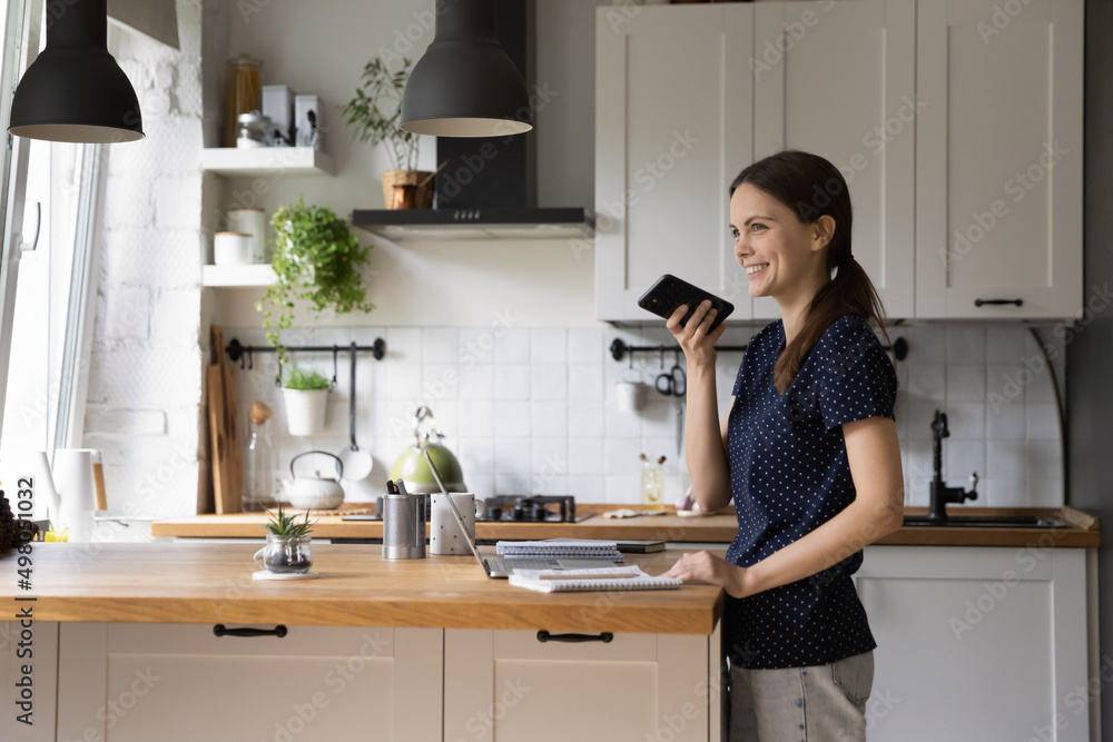 Young woman standing in domestic kitchen holding smart phone leaves voice message, share voicemail to friend, having pleasant conversation use speakerphone, easy convenient modern tech usage concept Young woman standing in domestic kitchen holding smart phone leaves voice message, share voicemail to friend, having pleasant conversation use speakerphone, easy convenient modern tech usage concept