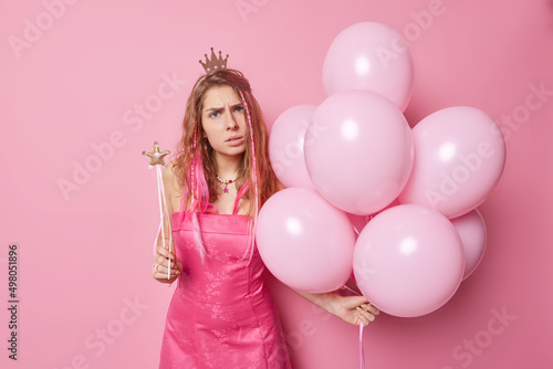 Indoor shot of displeased indignant European woman wears crown and dress comes on costume party holds bunch of inflated balloons and magic wand discontent to hear bad news. Special occasion.