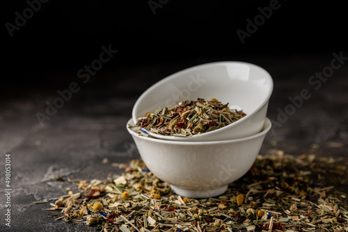 Herbal tea in a ceramic bowl and a glass jar on a black textural background. Detox and immunity tea. Herbal collection of chamomile, mint, lemon balm. rosehip and dried fruit pieces.