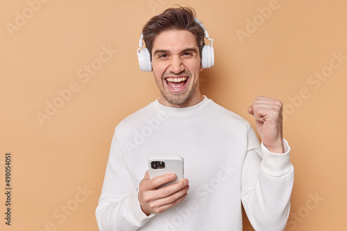 Successful positive man with dark hair clenches teeth rejoices good news wears casual white jumper uses smartphone for playing games online uses wireless headphones isolated over beige wall.