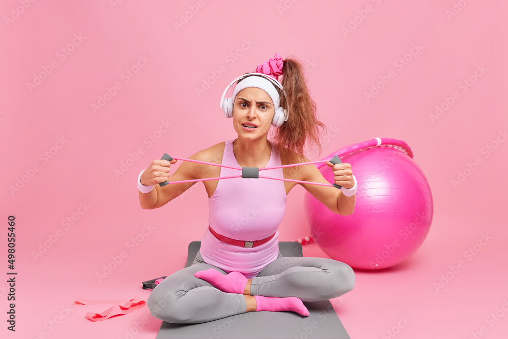 Serious woman with pony tail focused attentively at camera stretches resistance band wears sportsclothes sits in lotus pose on karemat surrounded by different sport equipment trains muscles. Serious woman with pony tail focused attentively at camera stretches resistance band wears sportsclothes sits in lotus pose on karemat surrounded by different sport equipment trains muscles.