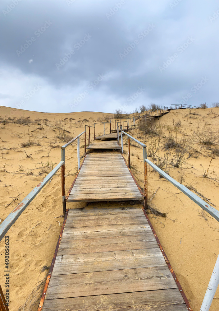 The staircase rises to a sandy dune. Climbing to the top in the desert. Sarykum dune. Dagestan, Russia. A unique sandy mountain in the Caucasus on a cloudy day. Grass grows on a sand dune.