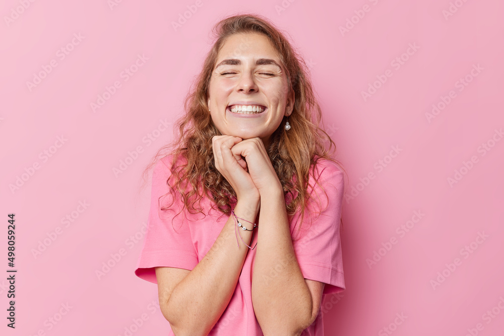 Positive young woman with long wavy hair keeps hands under chin closes eyes grins at camera recalls something pleasant wears casual t shirt isolated over pink background. Happy emotions concept Positive young woman with long wavy hair keeps hands under chin closes eyes grins at camera recalls something pleasant wears casual t shirt isolated over pink background. Happy emotions concept