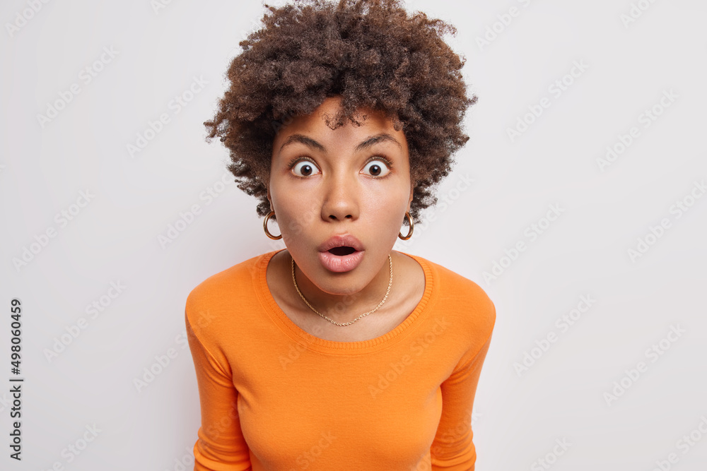 Indoor shot of stupefied beautiful woman with curly hair gasps amazed holds breath from amazement wears earrings casual orange jumper isolated over white background stares concerned at camera Indoor shot of stupefied beautiful woman with curly hair gasps amazed holds breath from amazement wears earrings casual orange jumper isolated over white background stares concerned at camera