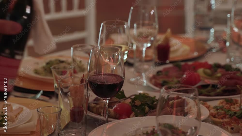Waiter pours red wine from bottle with napkin into clean wine glass. Glass stands on banquet table among variety of appetizer dishes, close-up. Festive banquet evening.