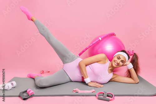 Fototapeta Naklejka Na Ścianę i Meble -  Cheerful sporty woman raises leg lies on fitness mat does aerobics wears headband and sportswear surrounded by sport equipment isolated over pink background. Body shaping training for weight loss