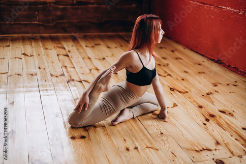Attractive athletic young woman with red hair doing yoga, stretching and training, in a sporty black bra and beige leggings, in the gym, studio with a ballet machine