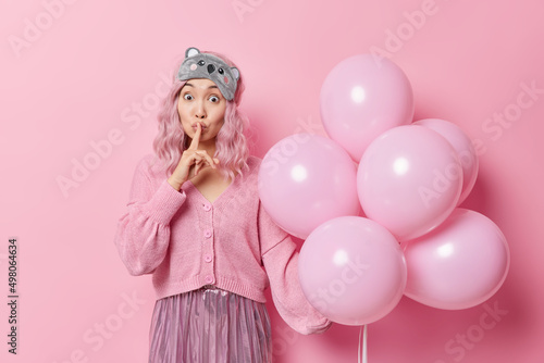 Mysterious surprised young Asian woman makes shush gesture keeps index finger over lips shares secret dressed in festive outfit holds bunch of balloons prepares surprise for friend on birthday