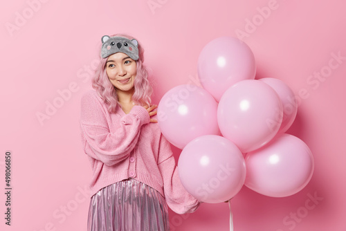 Dreamy lovely young Asian woman with long dyed hair dressed in festive clothes holds bunch of inflated balloons enjoys party time and holiday isolated over pink background. Holidays concept. © wayhome.studio 