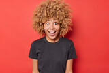 Blonde curly haired woman looks with happy surprised expression smiles broadly dressed in casual black t shirt isolated over bright red background reacts on unexpected offer. Human reactions