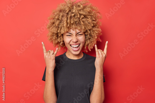 Positive young curly woman shows horn gesture enjoys rock music wears black casual t shirt isolated over vivid red background. Overjoyed curly haired female model demonstrates heavy metal sign