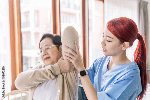 Nurse caregiver wearing scrubs exercises with a senior Asian woman to relieve a frozen shoulder of the senior patient with physiotherapy treatment. Home health care and nursing home concept.
