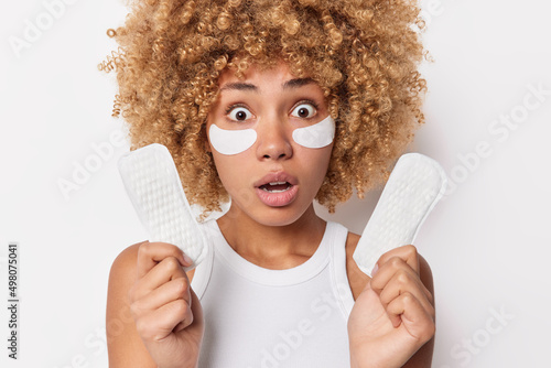 Shocked scared woman with blonde curly hair stares bugged eyes holds sanitary napkins applies beauty patches under eyes for skin treatment dressed in casual t shirt isolated over white wall. photo