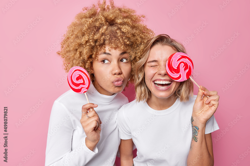 Φωτογραφία Two young women have fun pose with lollipops dressed in white clothes foolish around have sweet tooth isolated over pink background
