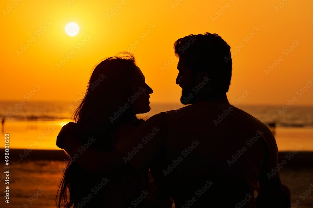 A beautiful silhouette of a young, cute tourist couple gazing with love, smiling, blushing on evening during sunset near beach on vacation or trip. 