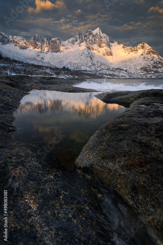 Beautiful sunrise on the rocky coast of the island of Senja in Northern Norway. The first warm sunlight shone very nicely on the snowy mountains, in the tide pool you can see the reflections.