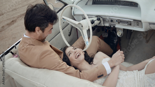 Happy smiling woman lying on the knees of her husband inside the cabriolet car. Action. Groom in suit holding hand of a beautiful bride.