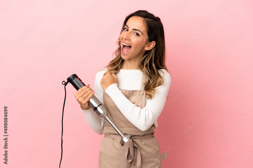 Young woman using hand blender over isolated pink background celebrating a victory