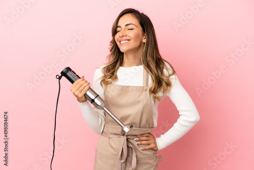 Young woman using hand blender over isolated pink background posing with arms at hip and smiling