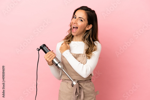 Young woman using hand blender over isolated pink background celebrating a victory