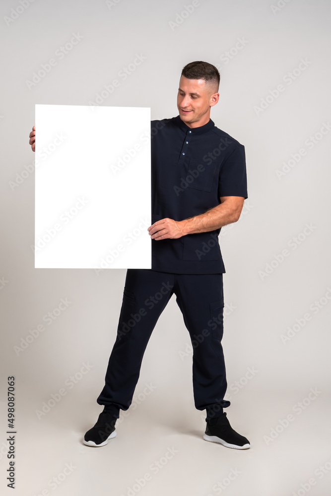 Blank poster in hand. Man with blank poster for an inscription. Banner in hands of man. He is showing a blank banner. Guy is standing full-length with a poster. Young guy on a gray background