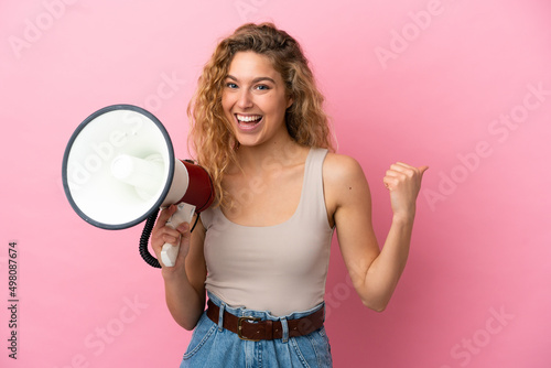 Young blonde woman isolated on pink background shouting through a megaphone and pointing side