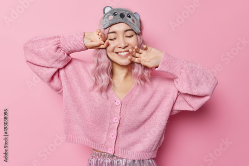 Satisfied Asian woman with dyed hair keeps eyes closed smiles gently imagines something pleasant wears sleepmask and jumper touches face gently isolated over pink background. Happy emotions concept