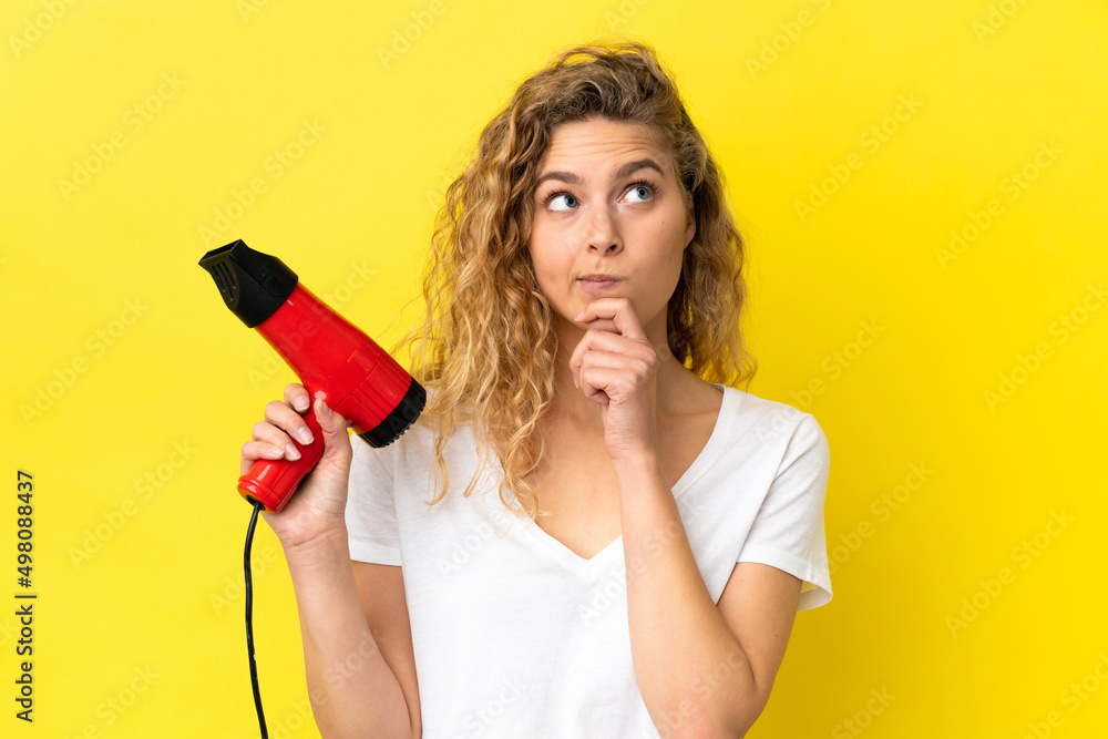 Young blonde woman holding a hairdryer isolated on yellow background having doubts and thinking Young blonde woman holding a hairdryer isolated on yellow background having doubts and thinking