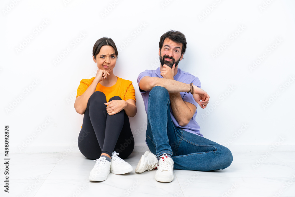 Young caucasian couple sitting on the floor isolated on white background having doubts and with confuse face expression Young caucasian couple sitting on the floor isolated on white background having doubts and with confuse face expression
