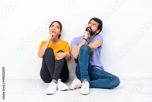 Young caucasian couple sitting on the floor isolated on white background standing and thinking an idea