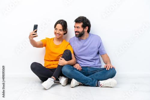 Young caucasian couple sitting on the floor isolated on white background making a selfie with the mobile