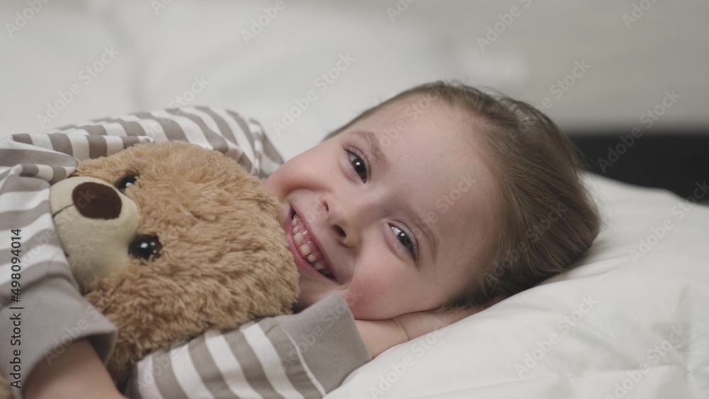 Happy girl smiles and hugs plush teddy bear lying on pillow