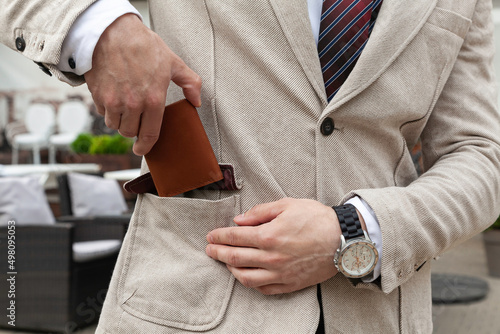 A stylishly dressed business man puts a brown leather wallet in his jacket pocket. Horizontal orientation, no face 