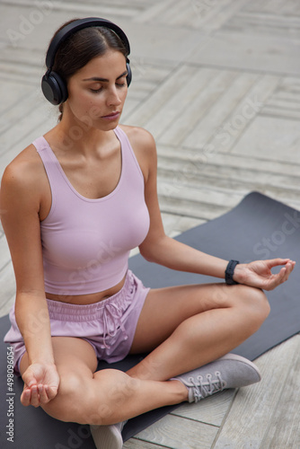 Vertcial shot of peaceful sporty young woman sits in lotus pose meditates on fitness mat breathes deeply keeps eyes closed listens relaxing music via headphones tries to find harmony. Keep calm