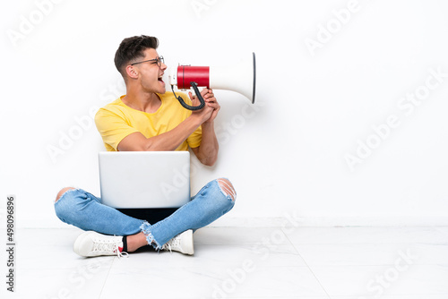 Young man sitting on the floor isolated on white background shouting through a megaphone