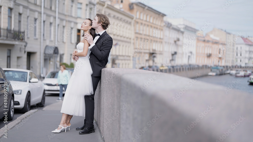 Side view of caring lovely just married couple embracing by the granite sidewalk. Action. Elegant bride and groom outdoors in the street.