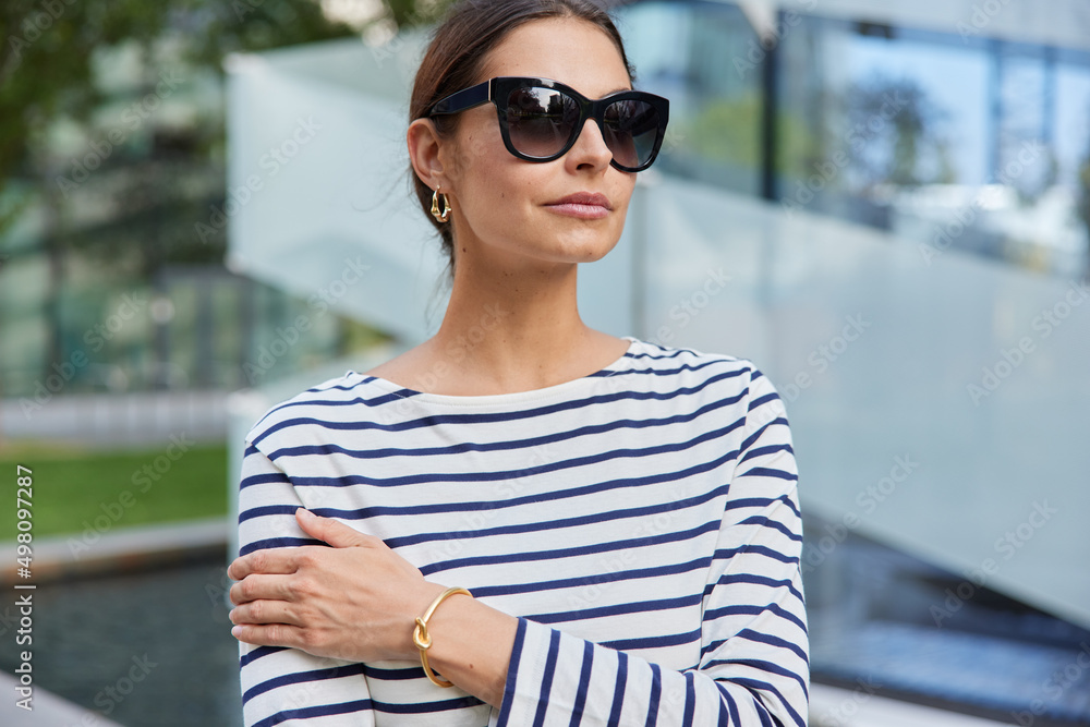 Horizontal shot of thoughtful European woman wears sunglasses casual striped jumper focused into distance walks outdoors poses at street against blurred background. Street lifestyle concept.