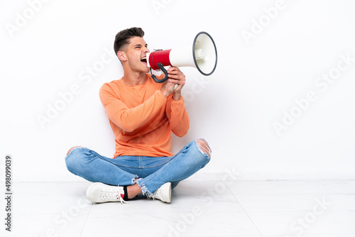 Young man sitting on the floor isolated on white background shouting through a megaphone
