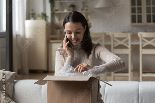Happy parcel receiver girl opening box, unwrapping parcel, talking to delivery service on mobile phone, ordering courier, giving feedback to internet shop, answering cellphone call