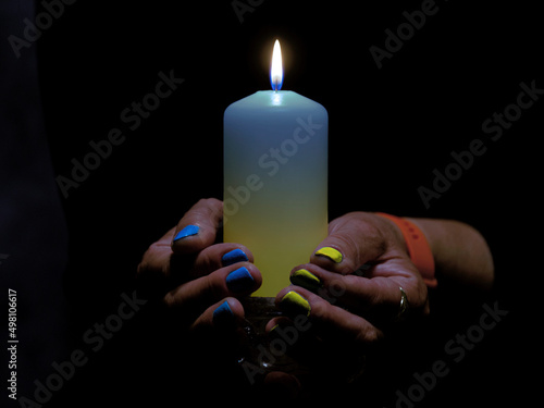 Płonąca świeca trzymana w kobiecych dłoniach. Świeca ma odcień niebieski i żółty. Na takie same kolory pomalowane są paznokcie. Jest to symbol solidarności z ofiarami wojny w Ukrainie, photo