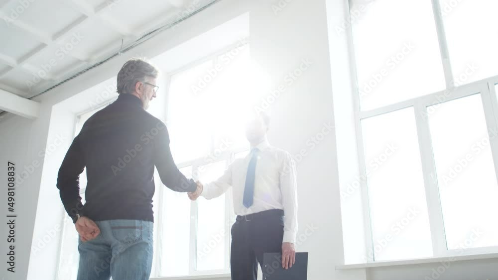 Realtor shakes hands with client after signing lease agreement, standing in front of window through which sun shines. Realtor and client shake hands after conclusion of contract Realtor shakes hands with client after signing lease agreement, standing in front of window through which sun shines. Realtor and client shake hands after conclusion of contract